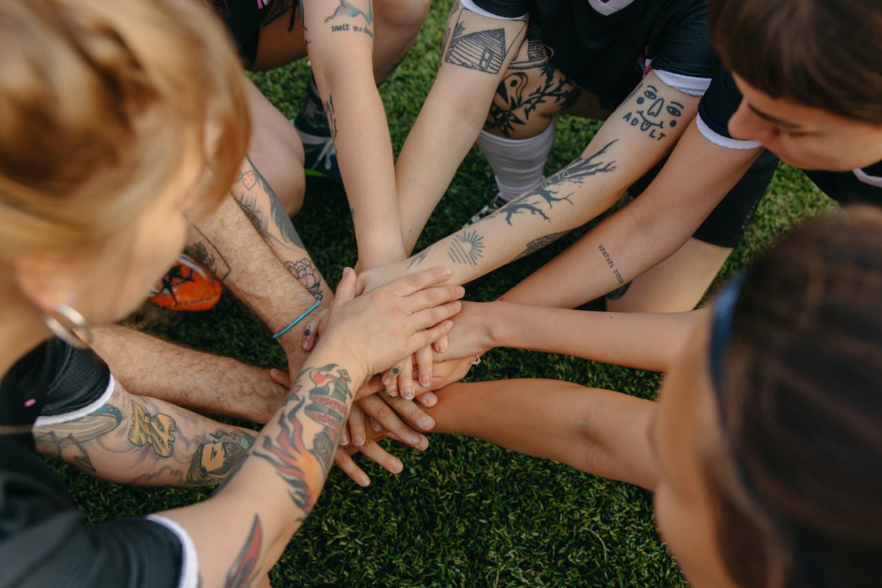 Tattooed team members join hands in a circle signifying unity and teamwork on a grassy field.